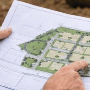 Especialista analisa com atenção a planta de um terreno que será dividido, segurando o projeto em primeiro plano, enquanto ao fundo é possível ver o próprio terreno em obras, com uma escavadeira em atividade, reforçando o contexto técnico e planejado do desmembramento