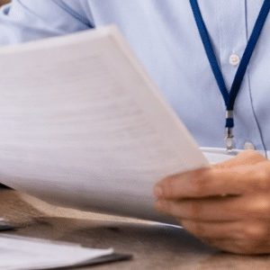 Um funcionário de cartório analisa atentamente documentos sobre a mesa, com expressão concentrada. Ao lado, há uma maquete detalhada de uma casa, reforçando o contexto imobiliário, em um ambiente interno bem iluminado e com aparência profissional.