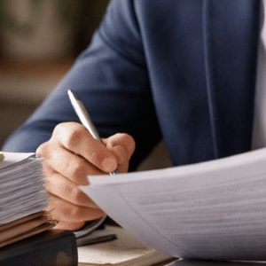 Advogado analisando documentos em sua mesa de escritório, com expressão concentrada, ao lado de uma pilha de papéis contendo uma maquete de casa sobreposta, em ambiente profissional com iluminação natural e composição realista institucional.