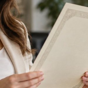 Mulher sorridente segurando a escritura de um imóvel em um escritório, com um profissional ao fundo e uma maquete de casa sobre a mesa.